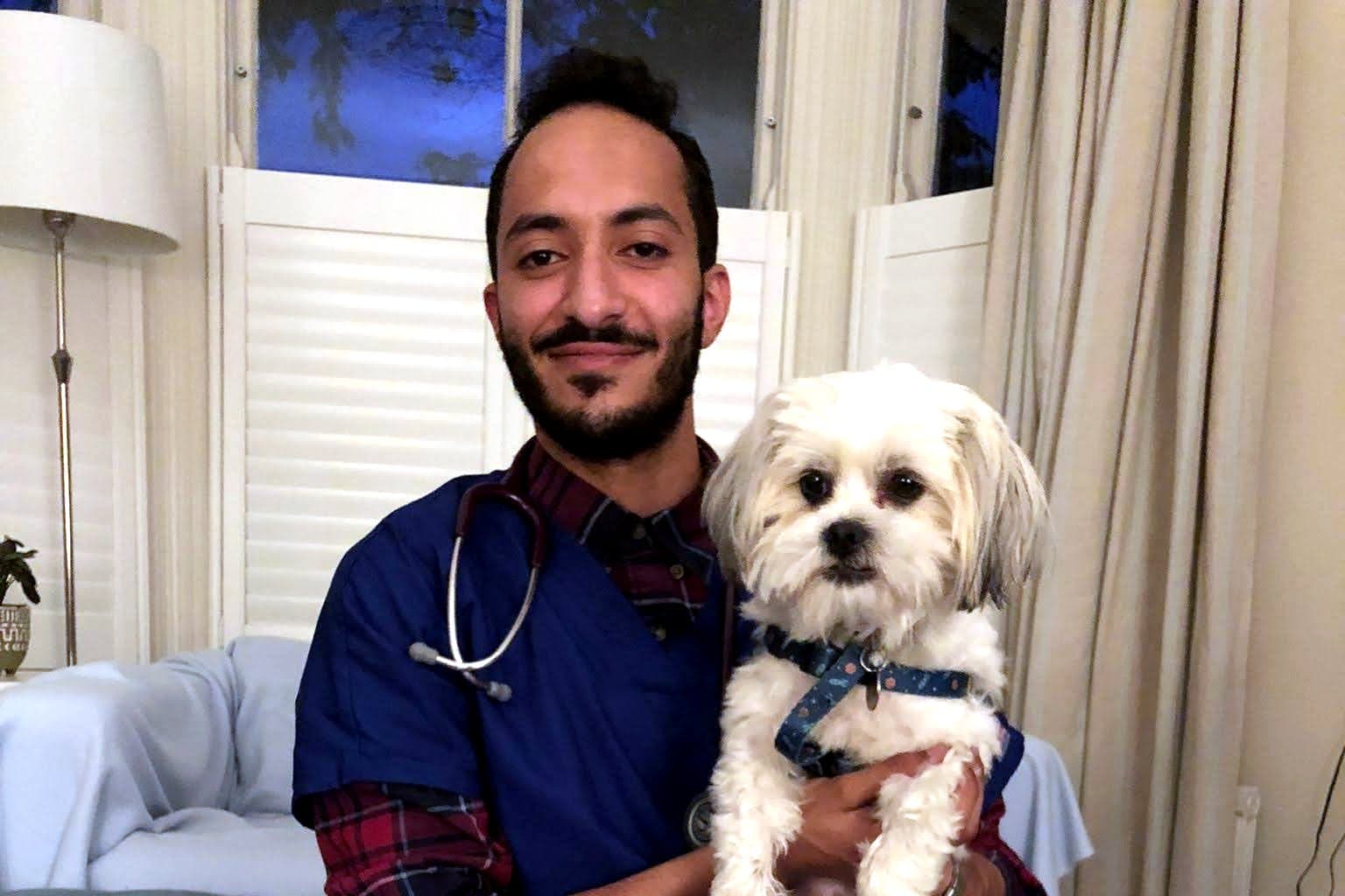 Ziad in scrubs and a stethoscope, smiling while holding a small white dog during a home visit