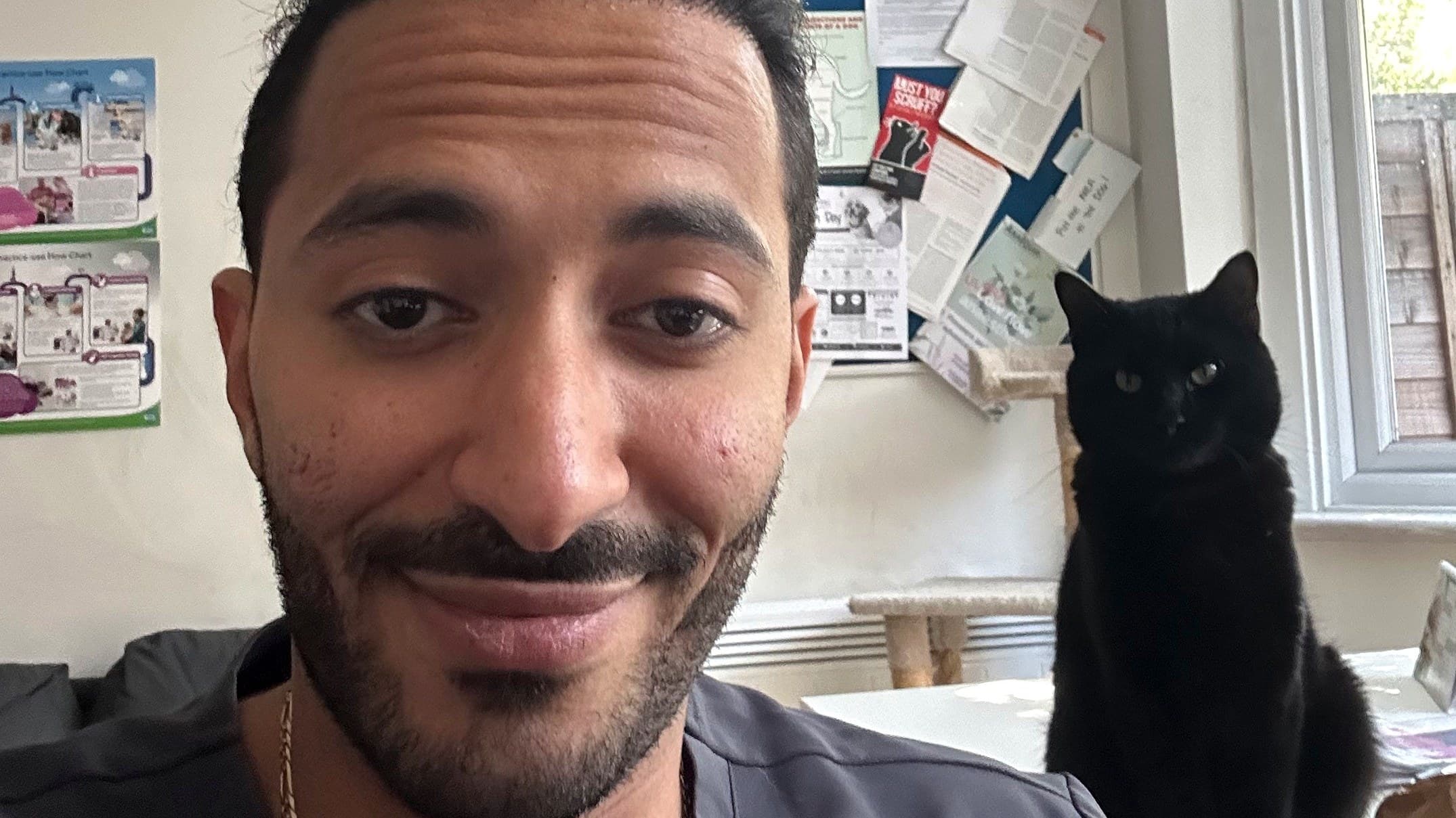 Ziad smiling at the camera in a treatment room, with a black cat sitting on the windowsill behind him